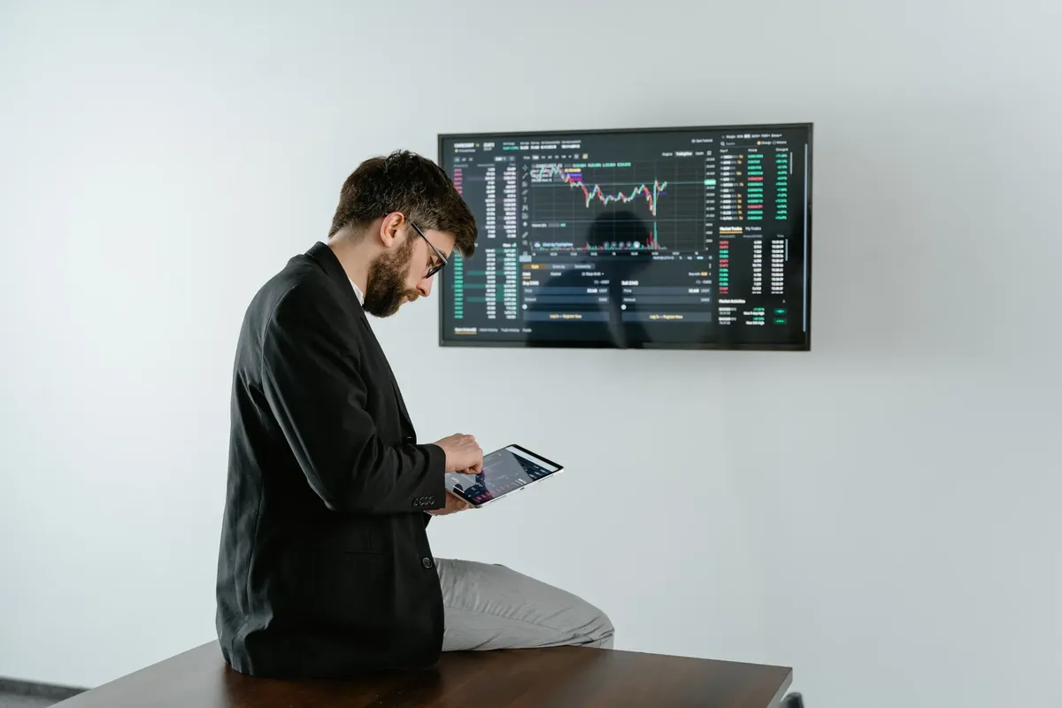 Young man in office analyzing financial graphs on a tablet with stock market screen displayed.