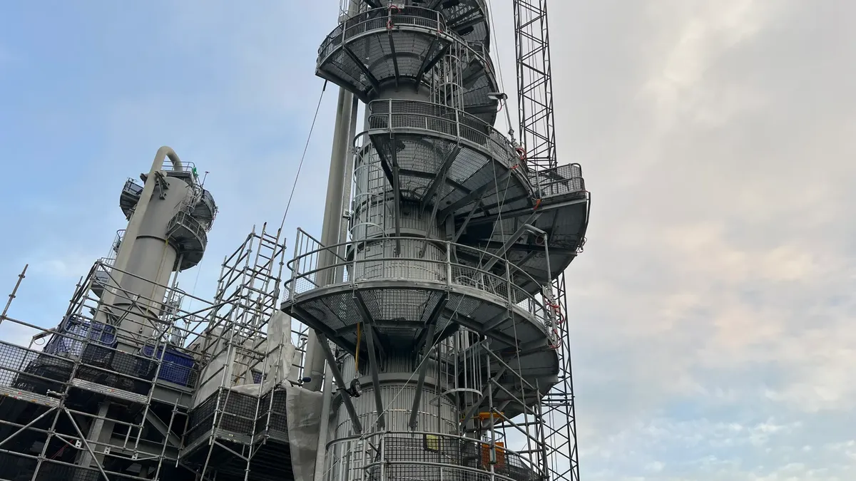View of an industrial refinery tower under construction with scaffolding against a cloudy sky.